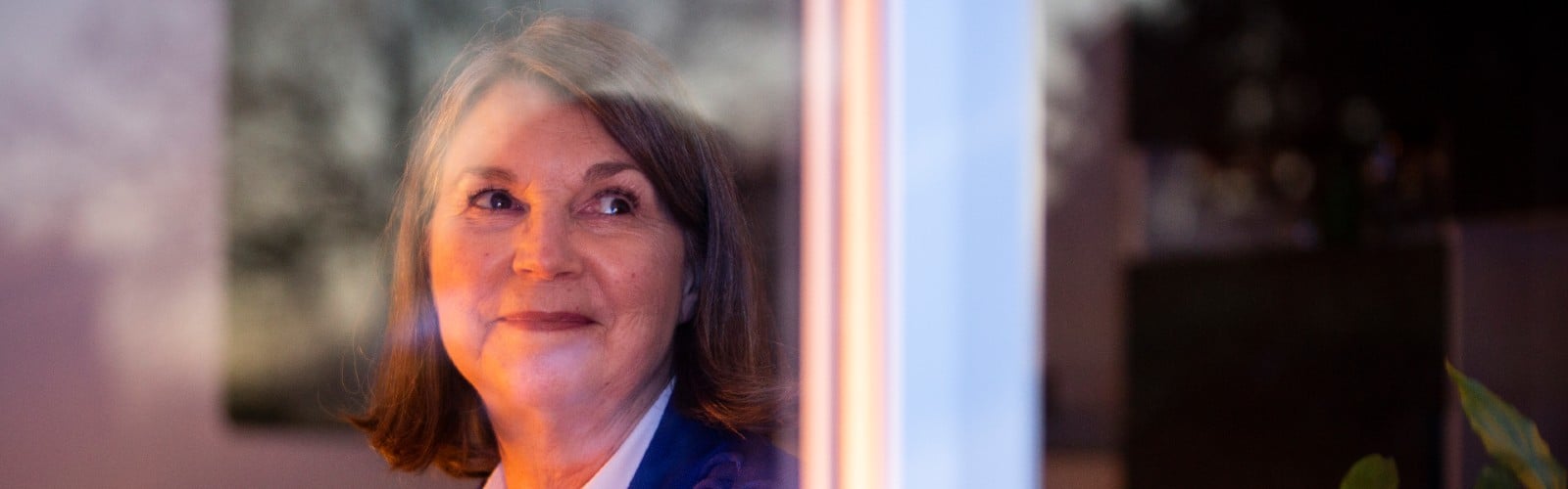 A middle-aged woman with short brown hair sits holding a cup of tea and looking out of her window, smiling slightly