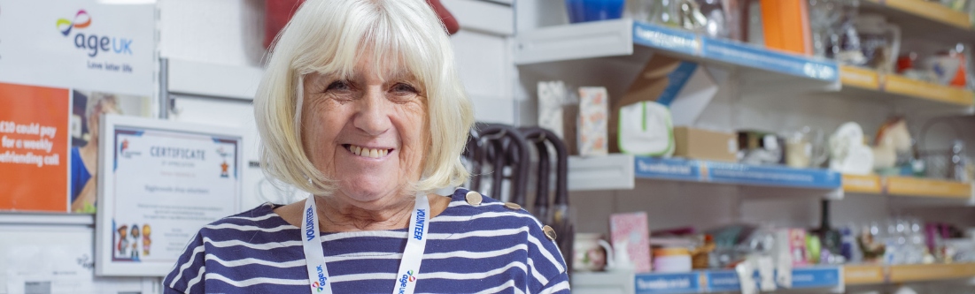 An older lady with grey hair, with an 'Age UK Volunteer' tabard, smiles from behind the desk of a charity shop