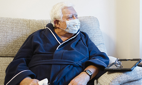 An older man sat on a sofa, wearing a face mask