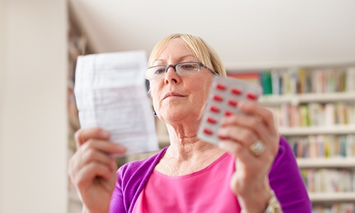 A woman with glasses reads her prescription, with a packet of pills in her other hand