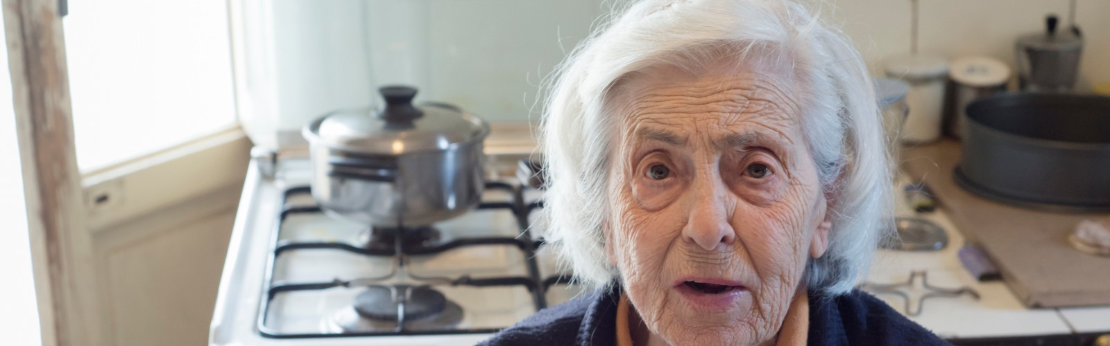 An older woman sits in her kitchen, looking concerned