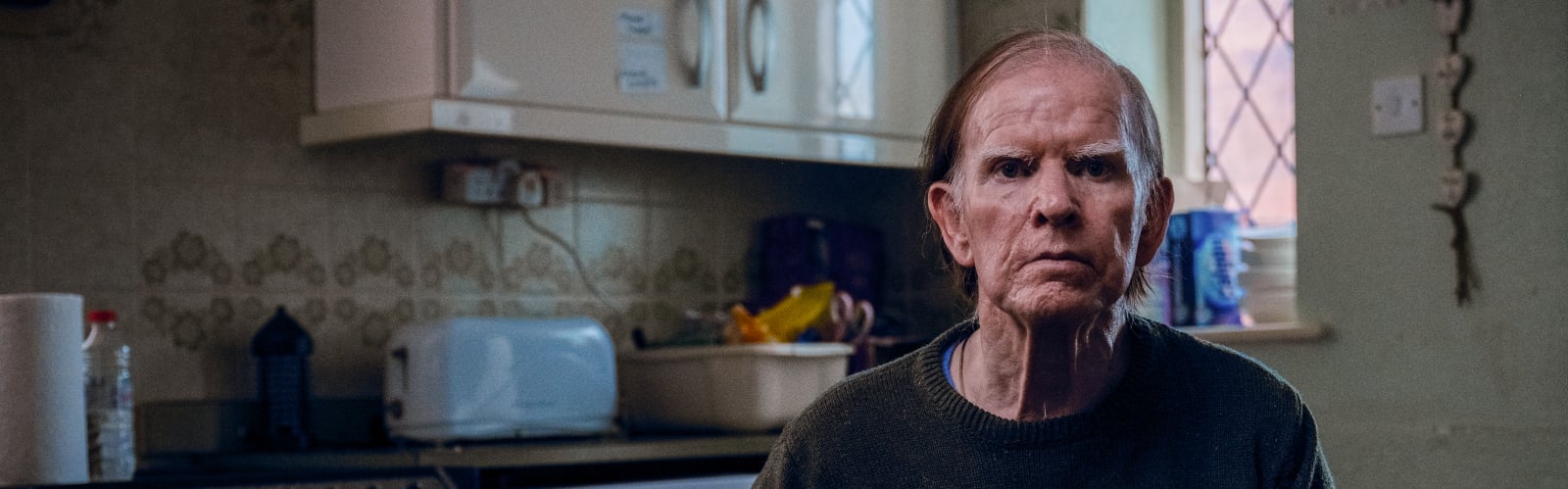 An older, slim man sits in his cold-looking kitchen