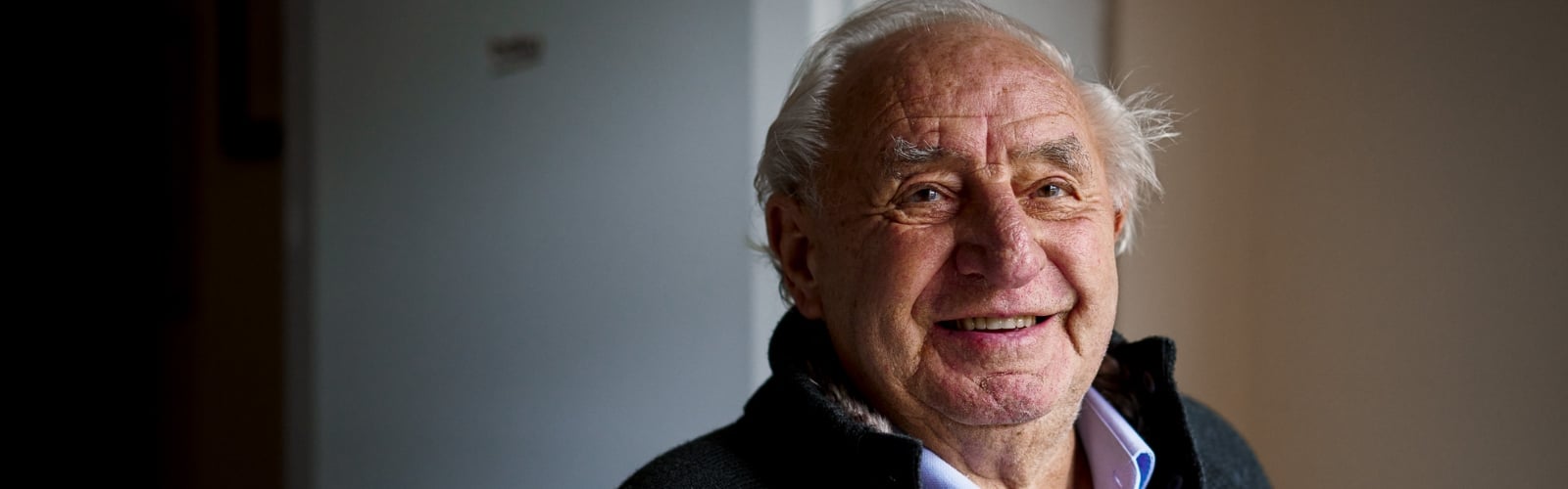 An older man smiles at the camera, stood next to a window