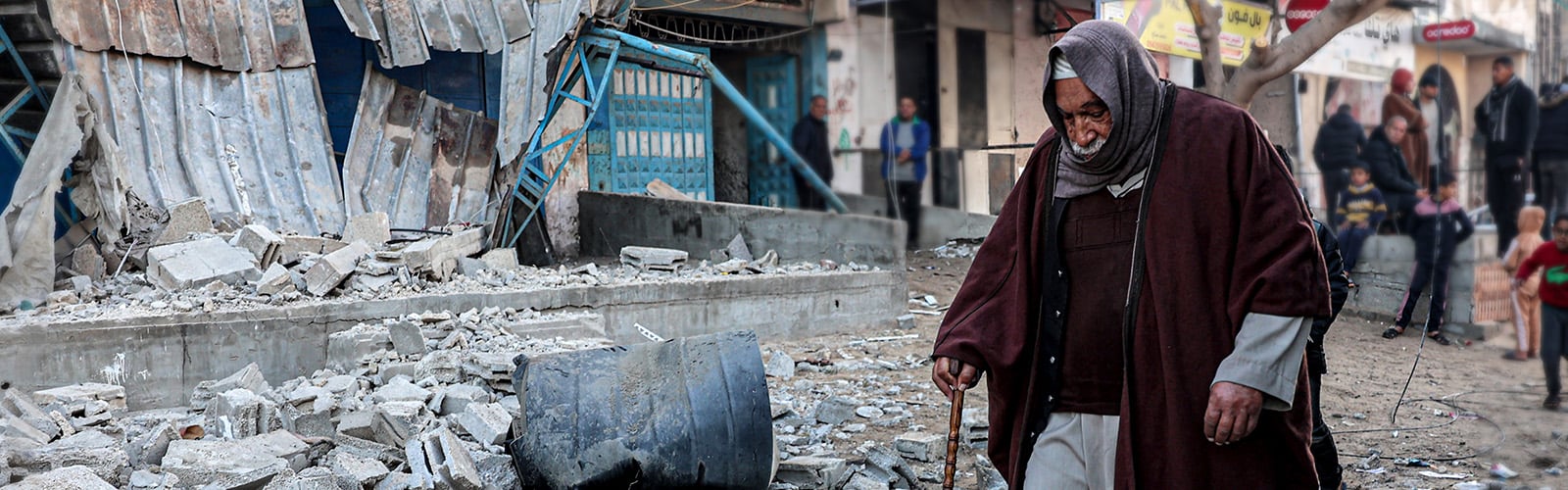 An older man on a stick, walking through rubble