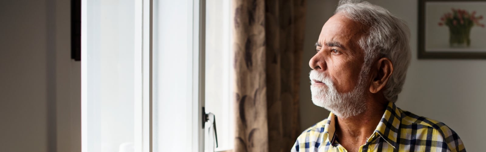 An older Asian man stands at the window, looking out of it sadly