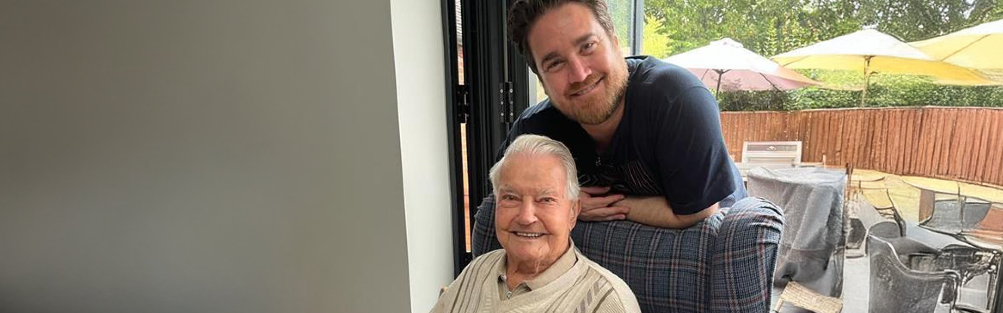 A middle aged man stands behind his grandfather, who sits in a chair. Both smile at the camera