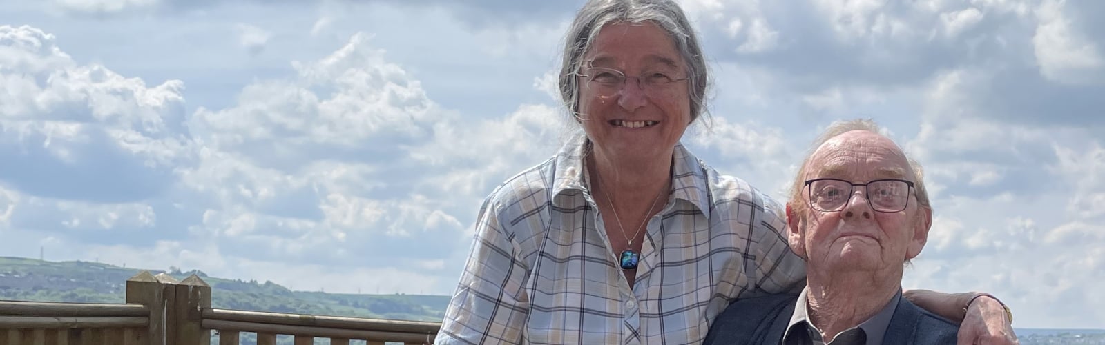 An older lady with glasses smiles to camera, with her arm around her husband, who sits in a wheelchair