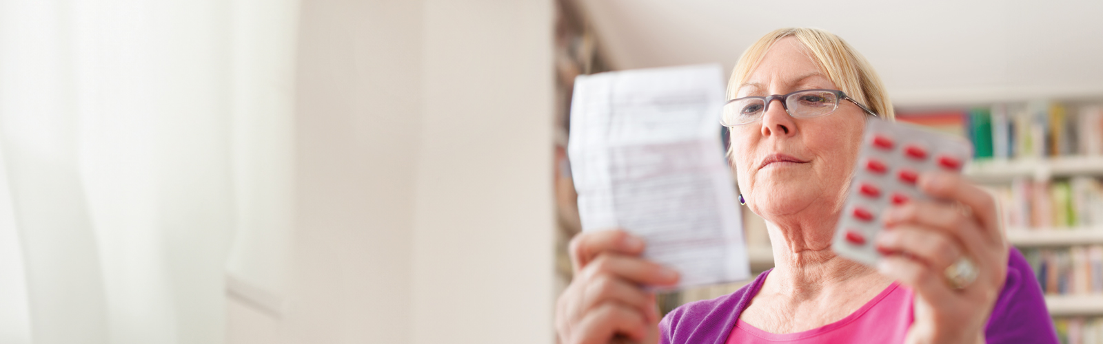 A woman with glasses reads her prescription, with a packet of pills in her other hand