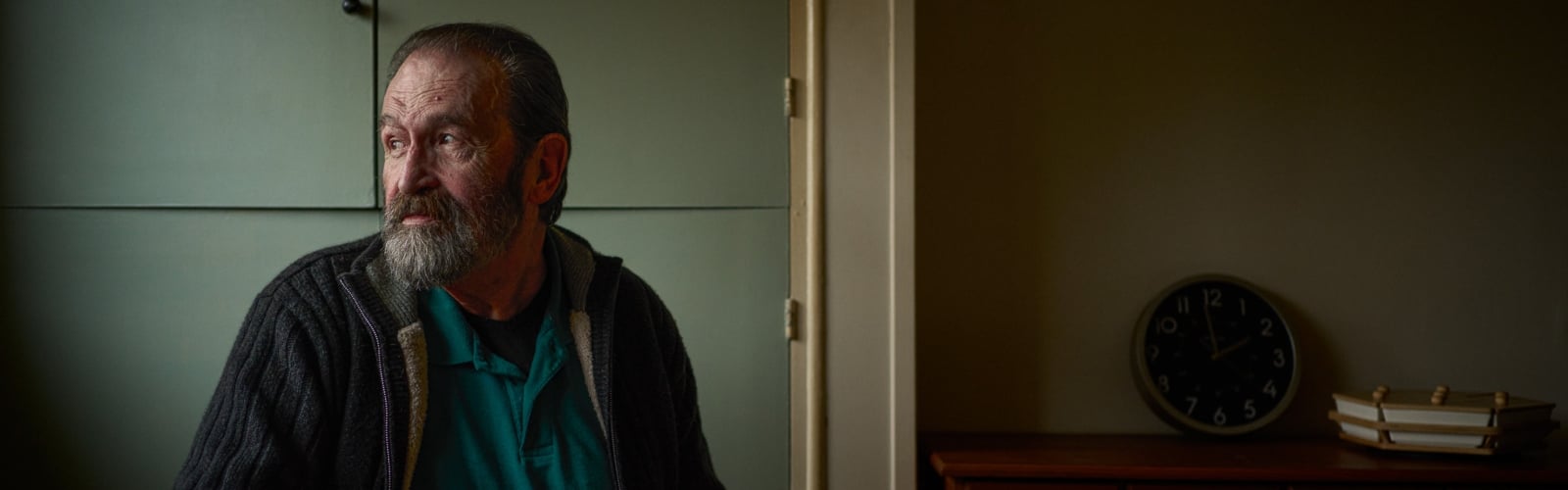 An older man with a beard sits in a dark room, looking out of the window