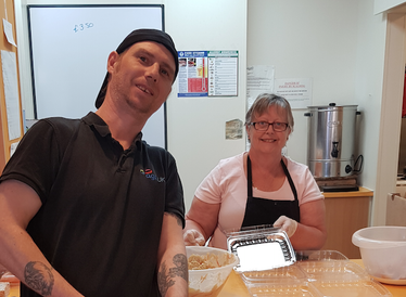 A man with a backwards facing baseball cap poses with a lady with glasses wearing an apron