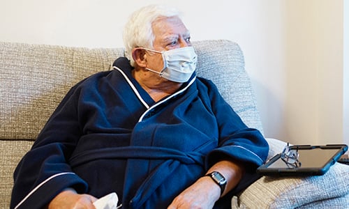 An older man, sat on a sofa and wearing a protective face mask