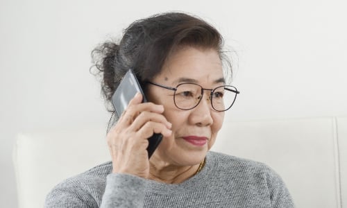 An older Asian woman with glasses talks on the phone, looking concerned