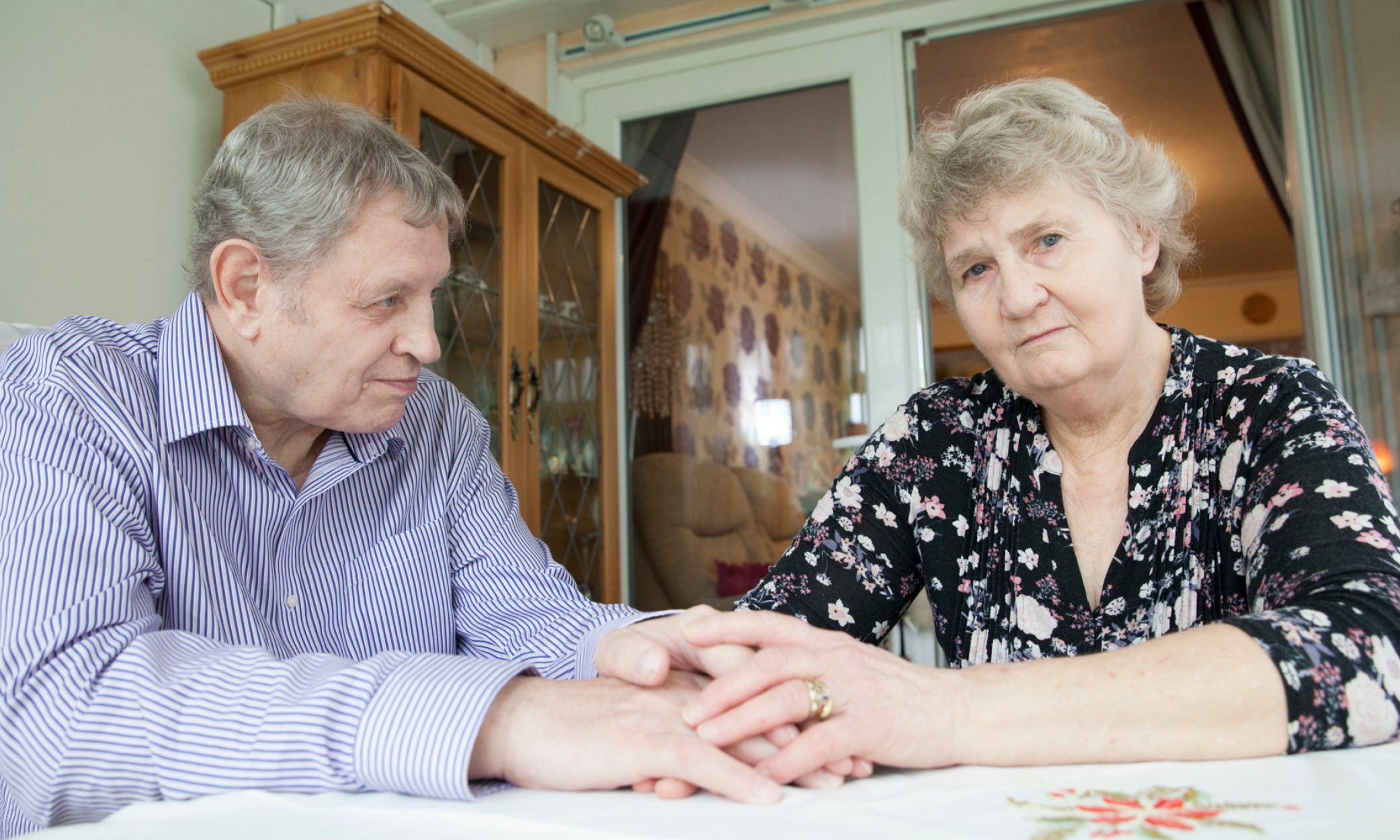 Elaine and Michael, a married couple, sit at a table, holding hands