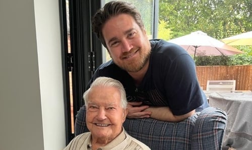 A middle aged man stands behind his grandfather, who sits in a chair. Both smile at the camera