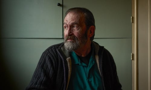 An older man with a beard sits in a dark room, looking out of the window