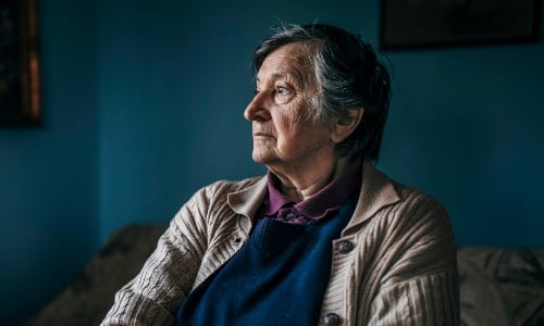 An older woman in a thick cardigan sits in a dark, cold-looking room, staring thoughtfully out of the window